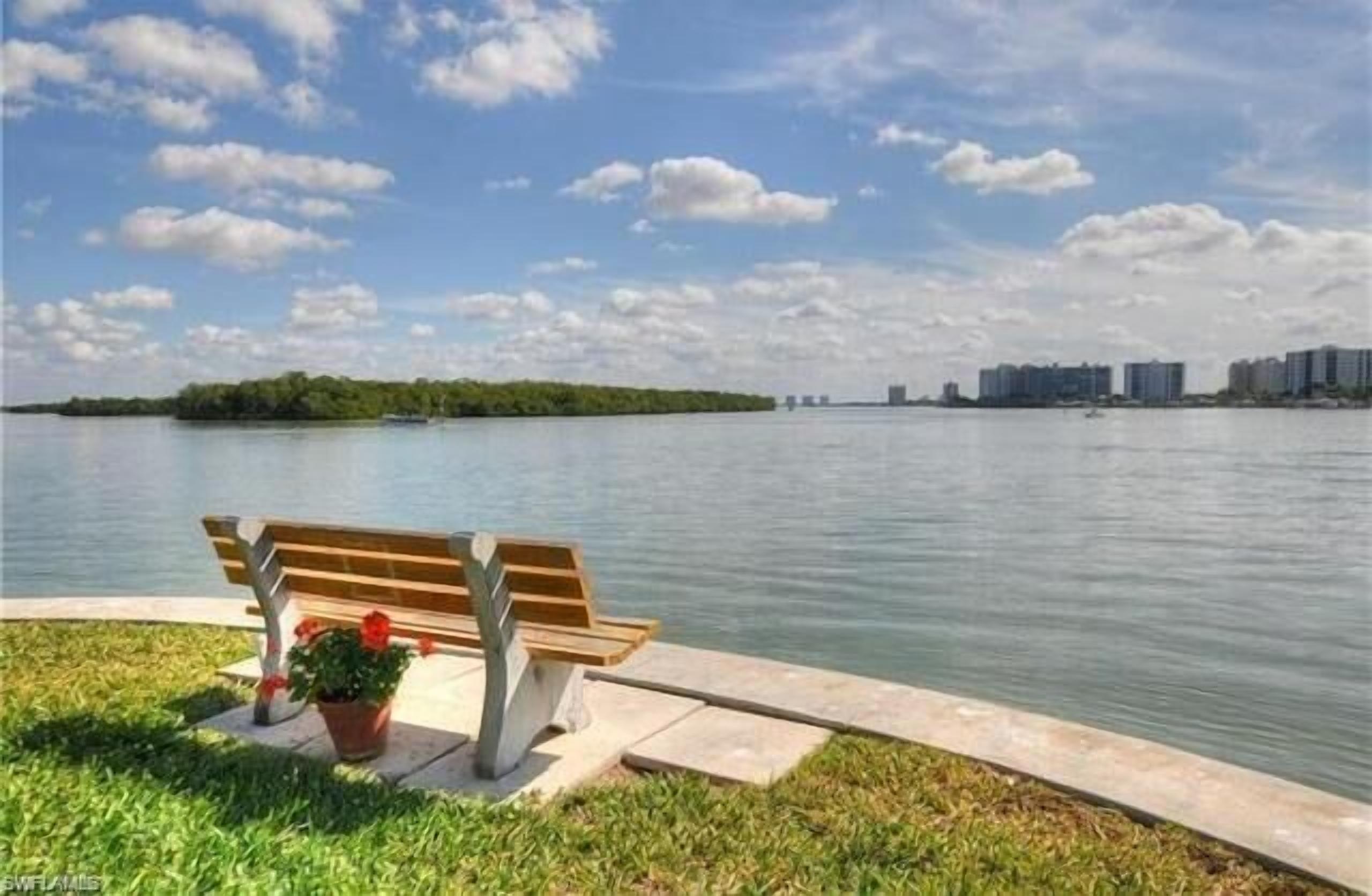A bench by a calm body of water with a distant view of buildings.
