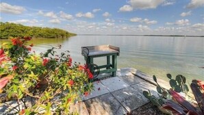 A serene lakeside setting with a small table and chair, surrounded by lush greenery and flowering plants. - ✨Amazing Bay Views + Amenities-Windward Point 507✨ (Fort Myers Beach)