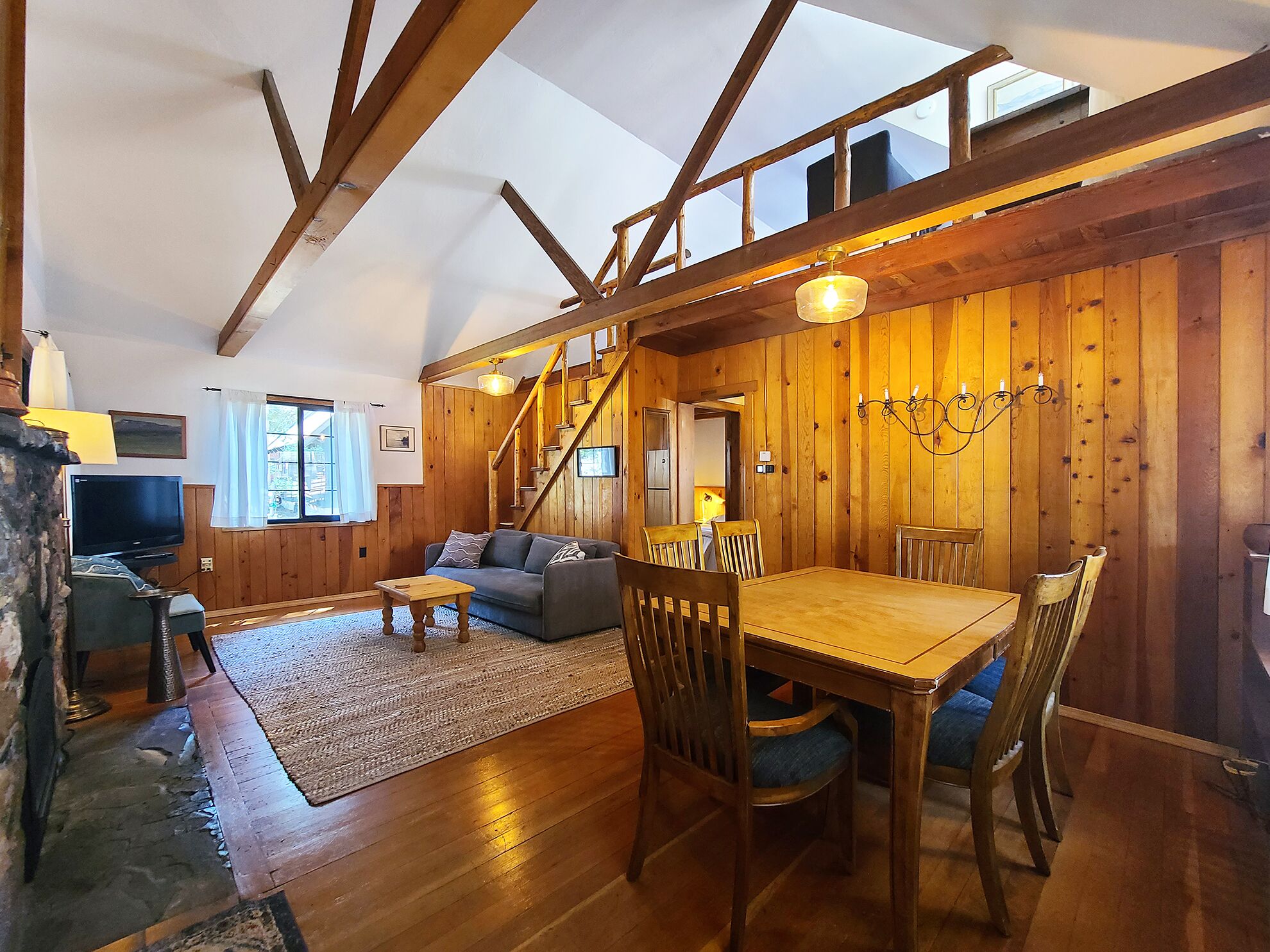 Ein gemütliches Blockhaus-Interieur mit einem Holzesstisch, Stühlen, einer Couch und einer Treppe, die in ein Obergeschoss führt.