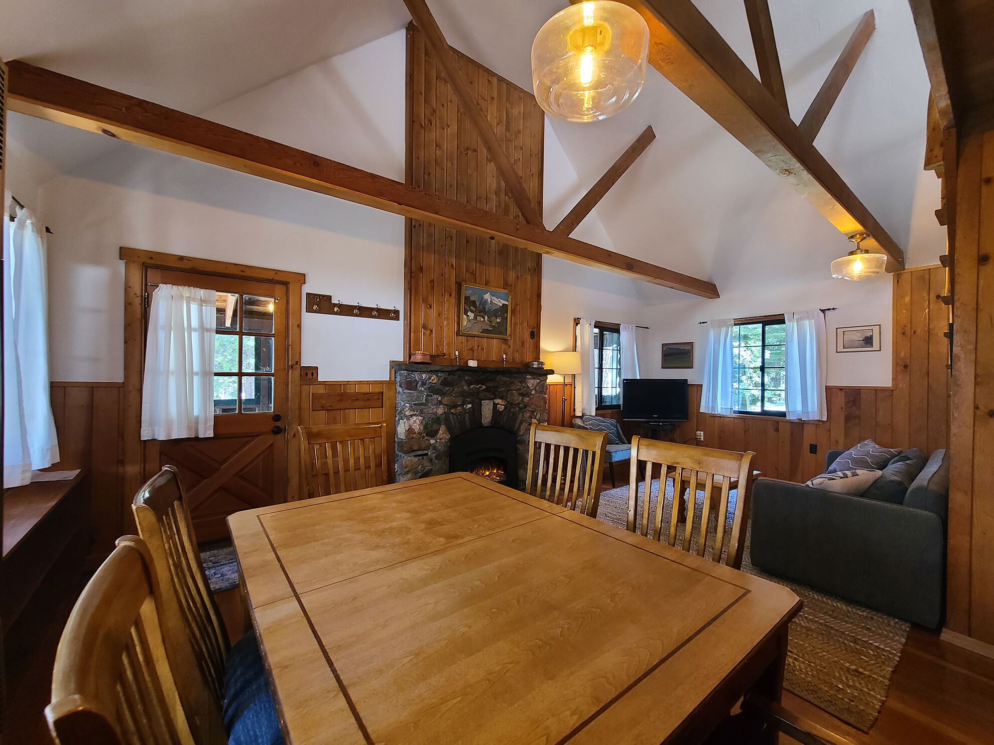 A cozy cabin interior with a wooden dining table, chairs, a stone fireplace, a television, and a couch.