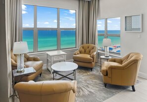 Ein Hotelzimmer mit großem Fenster und Meerblick, einer Couch, zwei Sesseln, einem kleinen Tisch und einer Lampe.
