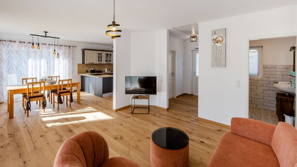Una sala de estar moderna con piso de madera, un comedor con mesa y sillas, una cocina al fondo y un televisor sobre un mueble.