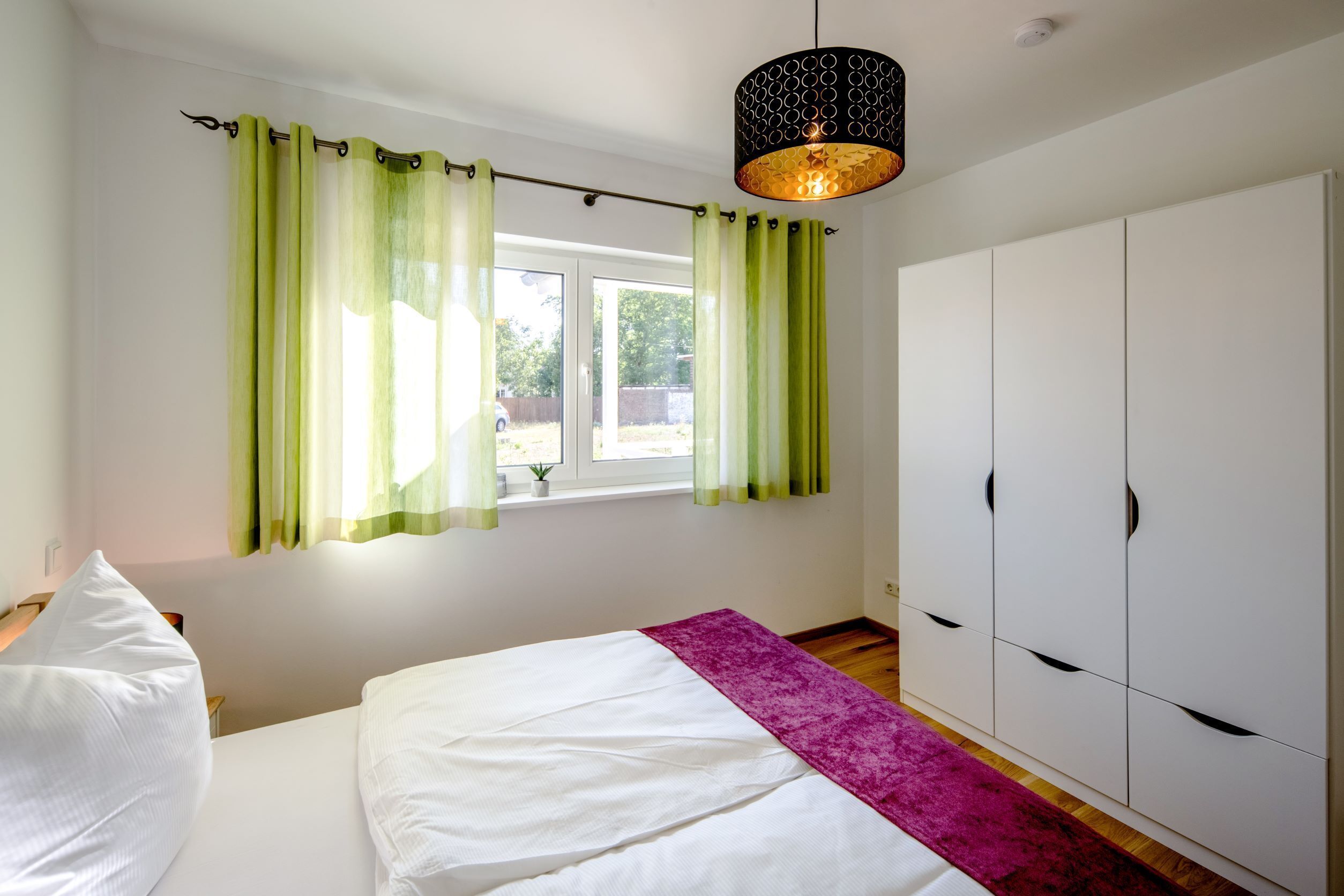 A bedroom with a bed, a wardrobe, a window with curtains, and a hanging light fixture.
