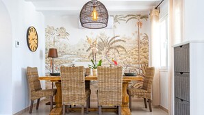A dining area with a wooden table and wicker chairs, a wall clock, and a mural of tropical scenery.