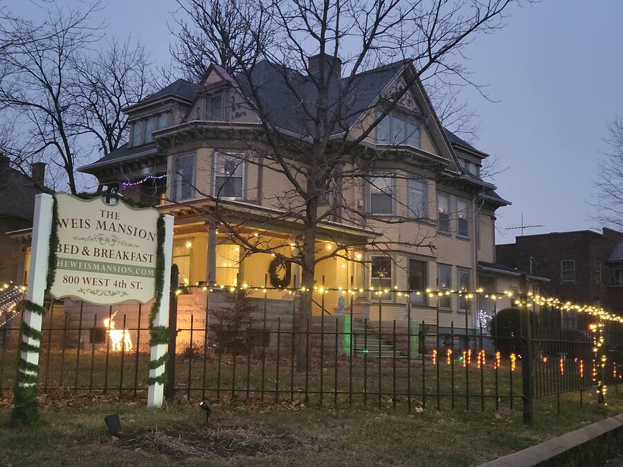 A large, historic house with a porch decorated with string lights and a sign advertising bed and breakfast services.