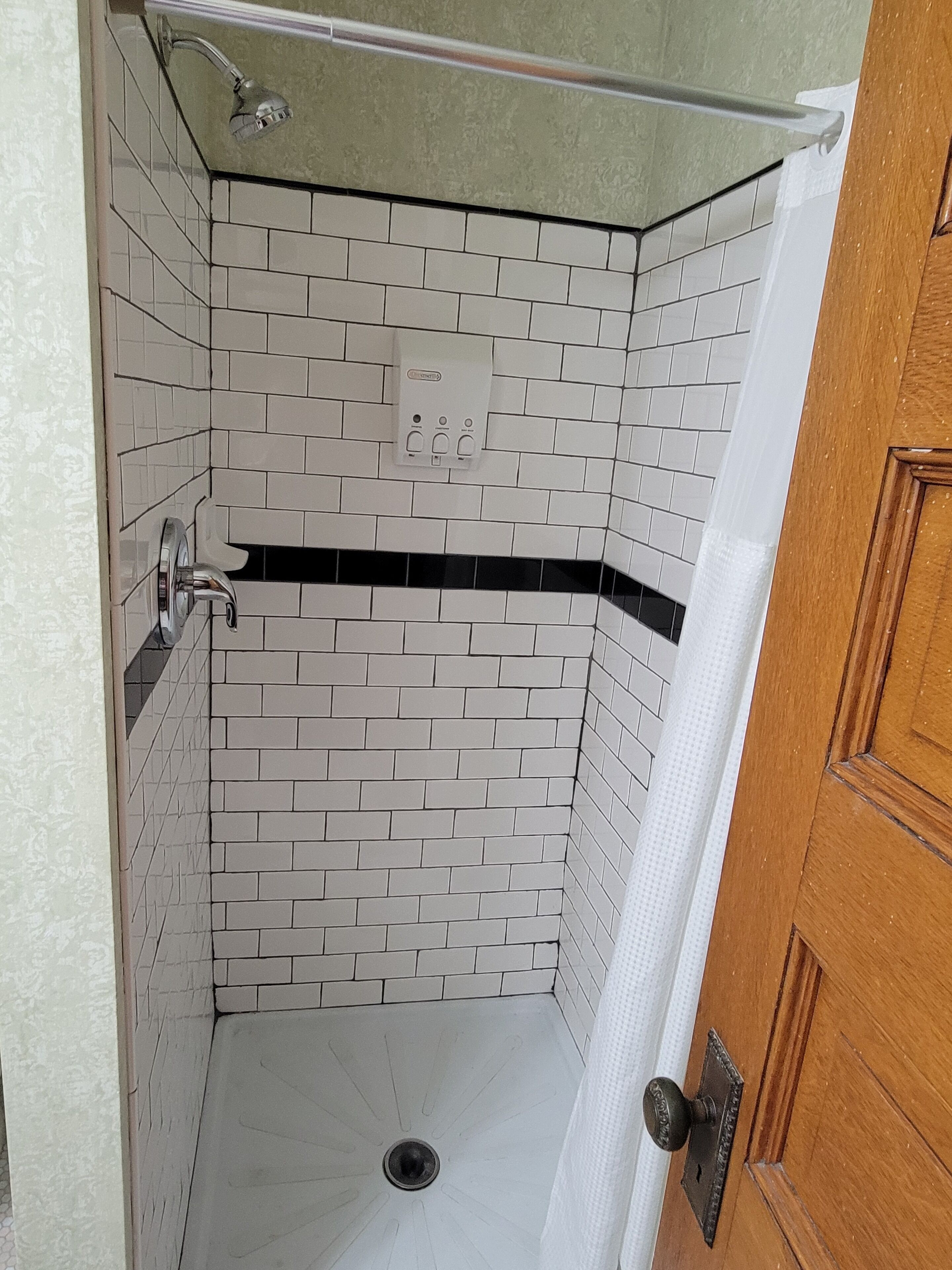 A small, enclosed shower area with white and black tiled walls, a showerhead, and a drain.