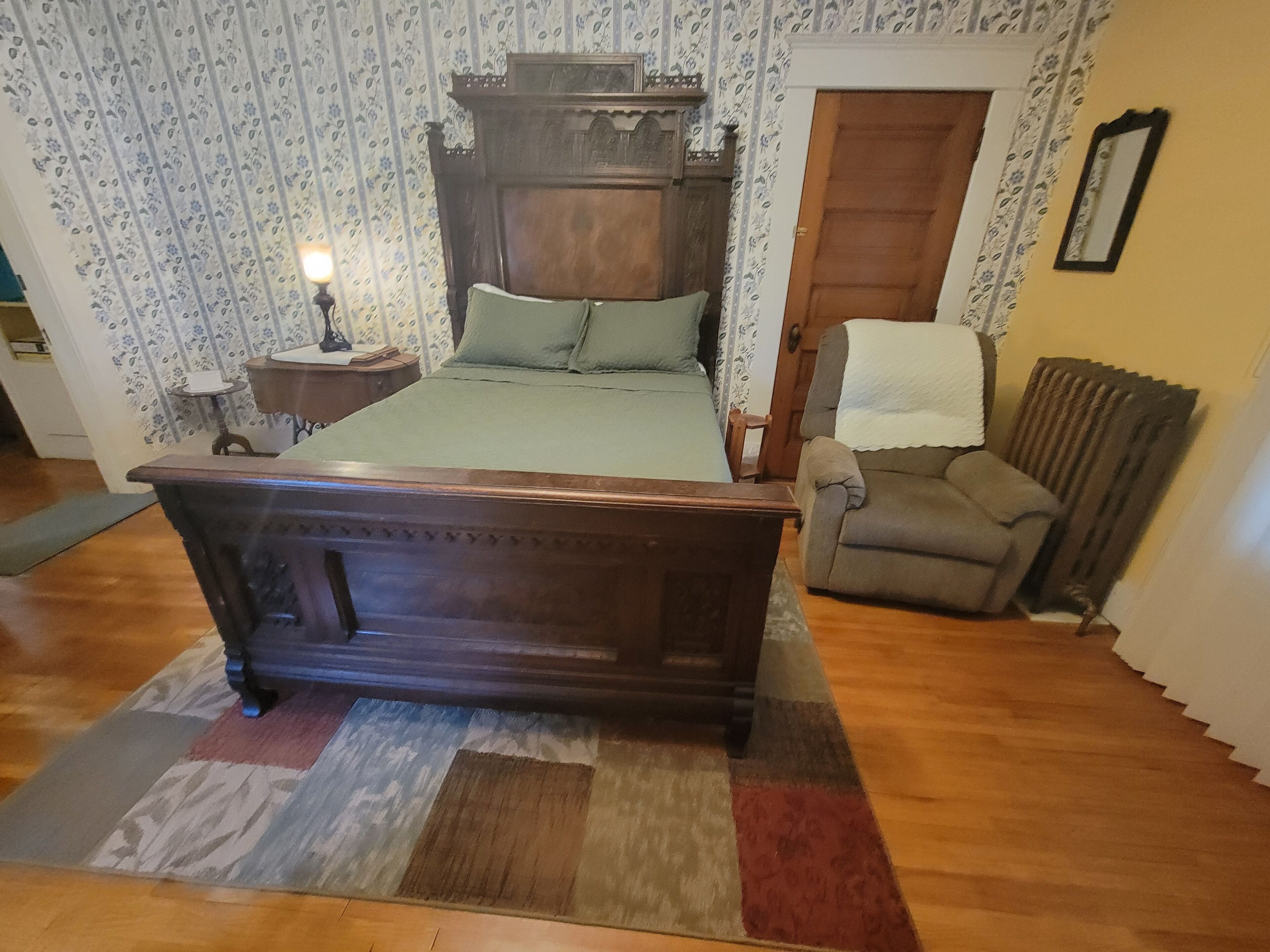 A bedroom with a four-poster bed, a wooden nightstand, a chair, a radiator, and patterned wallpaper.
