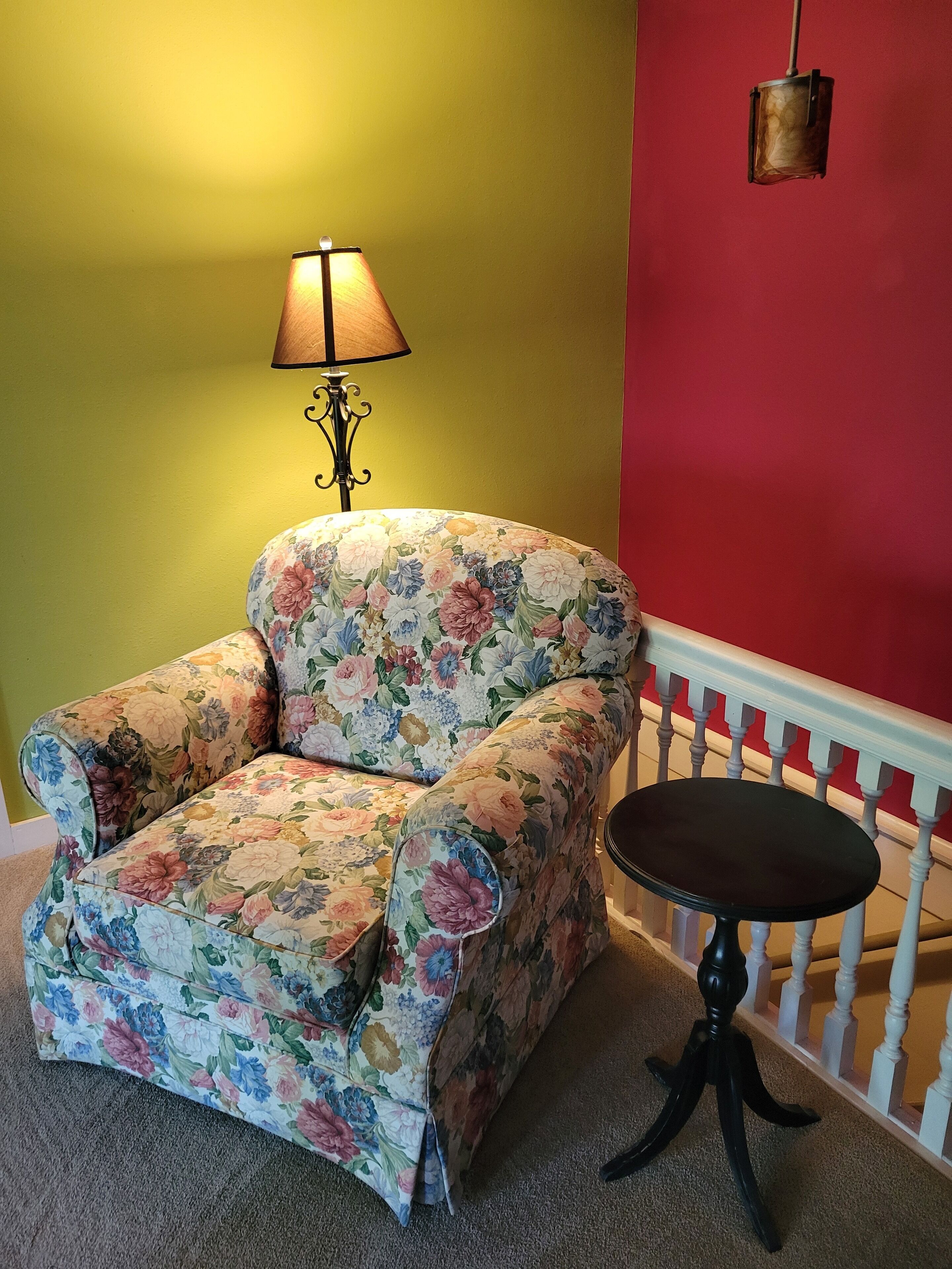 A floral armchair with a matching ottoman, a small round table, a lamp, and a red wall with a white railing.