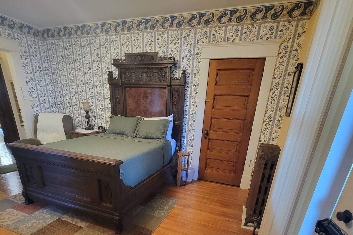 A bedroom with a wooden bed, a nightstand, a chair, a patterned wall, a wooden door, and a radiator.