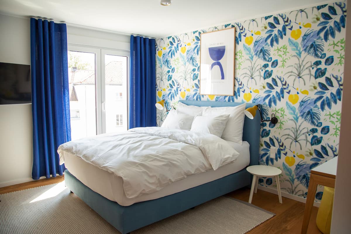 A bedroom with a bed, a small stool, a table, and a window with curtains.