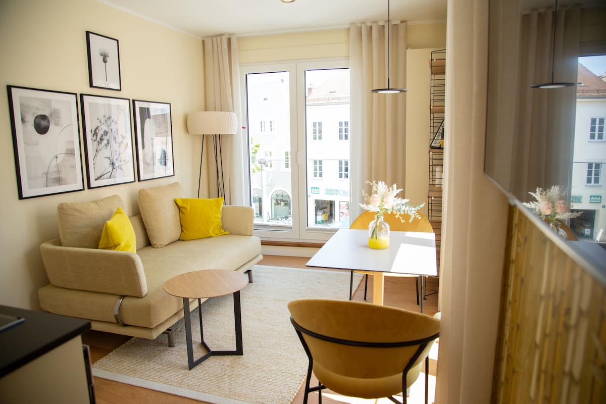 A modern living room with a sofa, a small round table, and a yellow chair. There are framed pictures on the wall and a window with curtains.