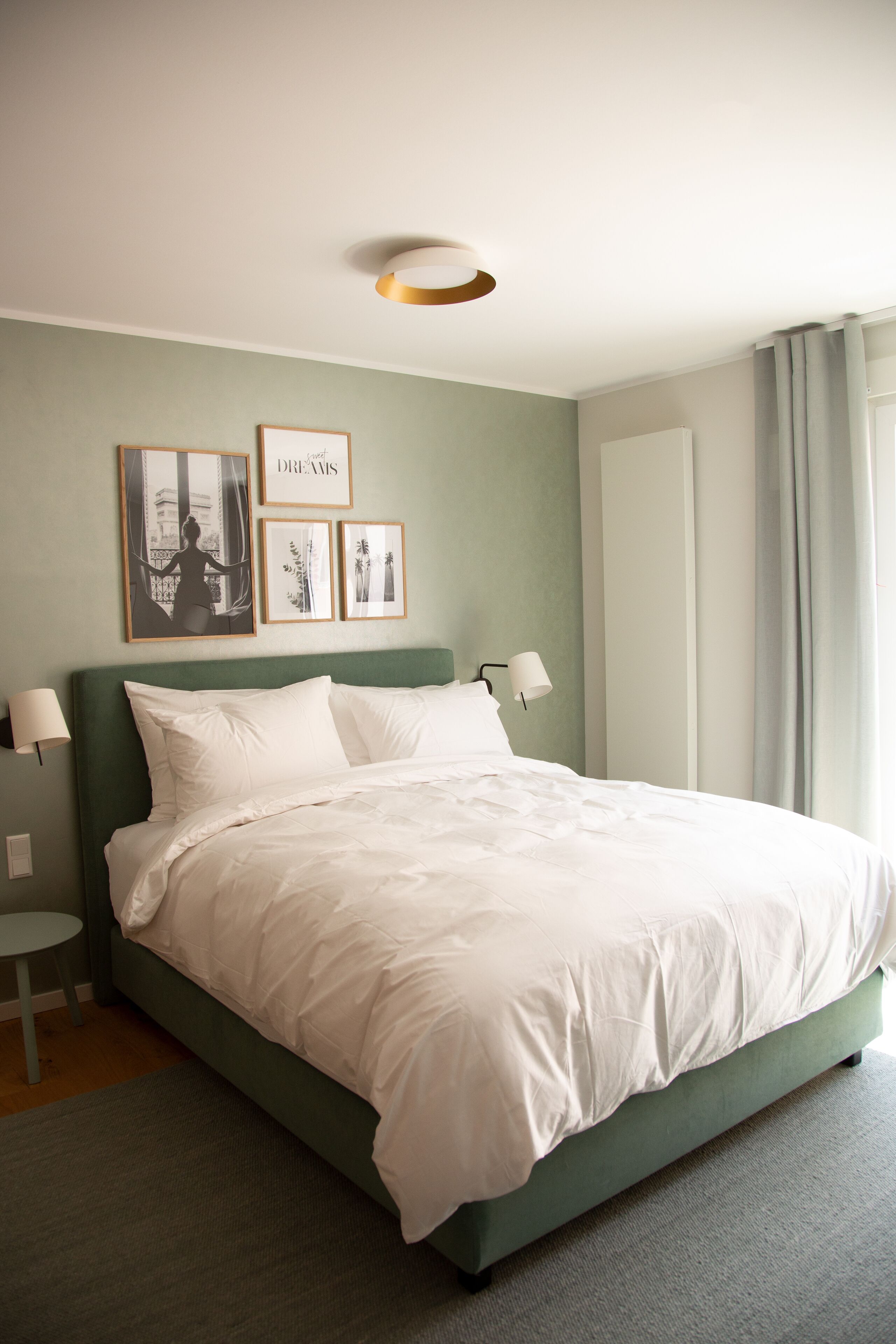 Ein ordentlich bezogenes Bett mit weißen Leinentüchern, ein grüner Kopfteil und eine Wandlampe daneben. Darüber an der Wand befinden sich gerahmte Bilder.