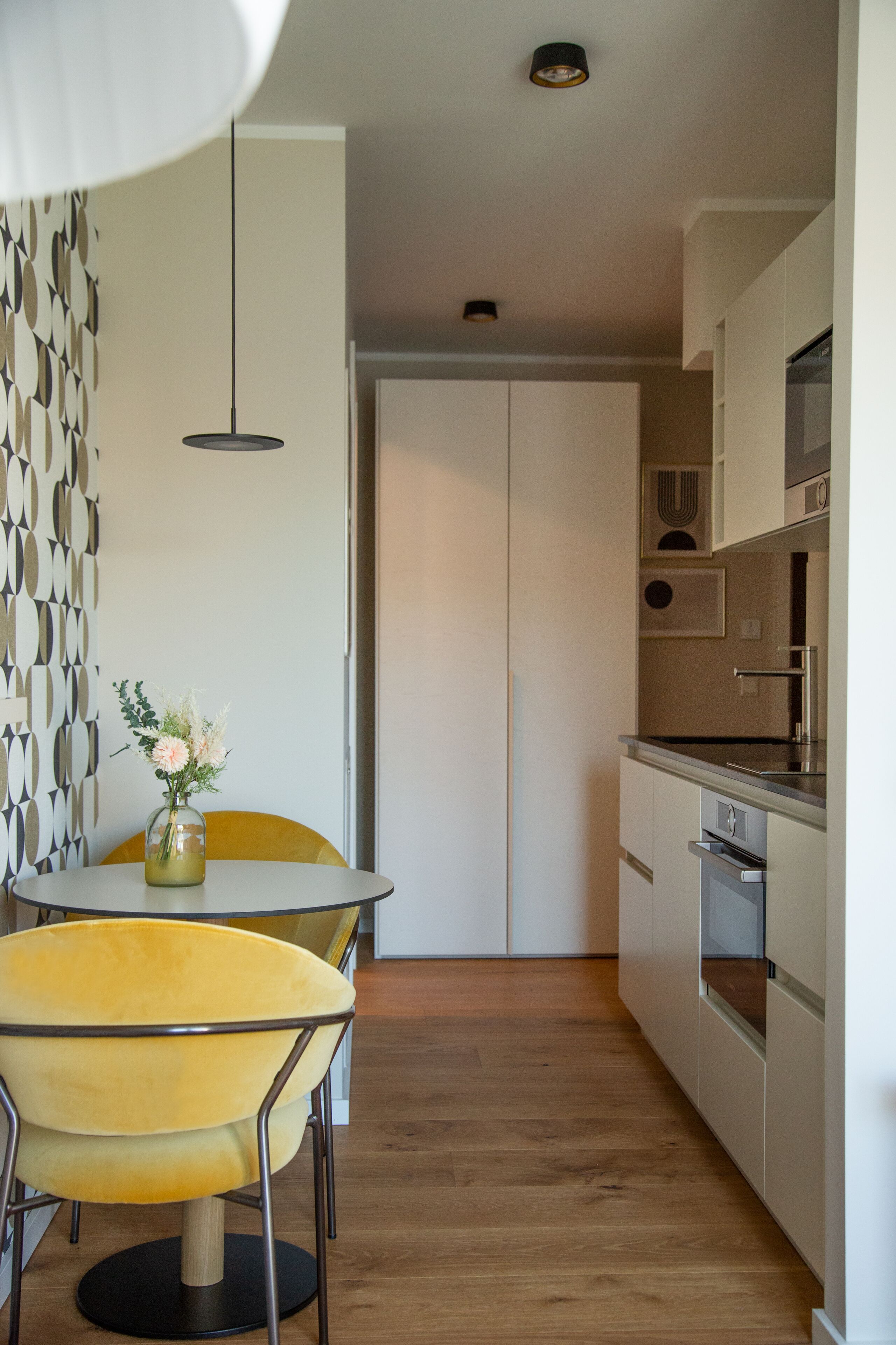 A modern kitchen with a dining area, featuring a yellow chair, a vase with flowers, and a patterned wall.