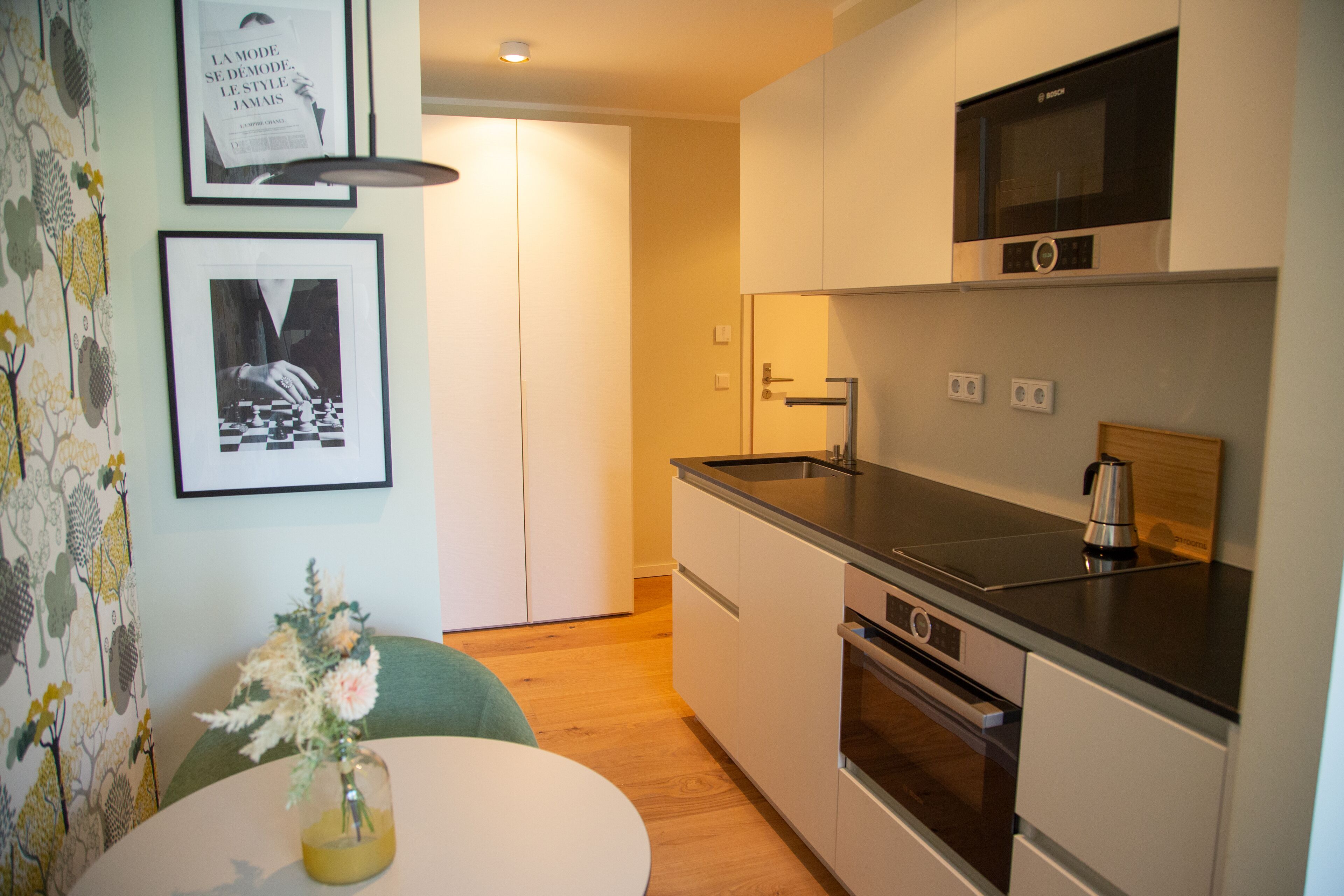 A compact kitchen with a microwave, oven, and sink. There is a framed picture on the wall and a vase with flowers on a table.