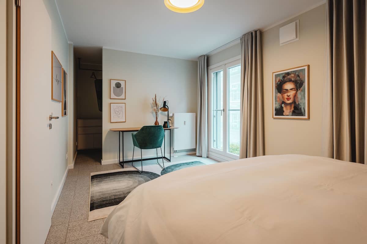 A hotel room with a bed, desk, chair, and a painting on the wall.