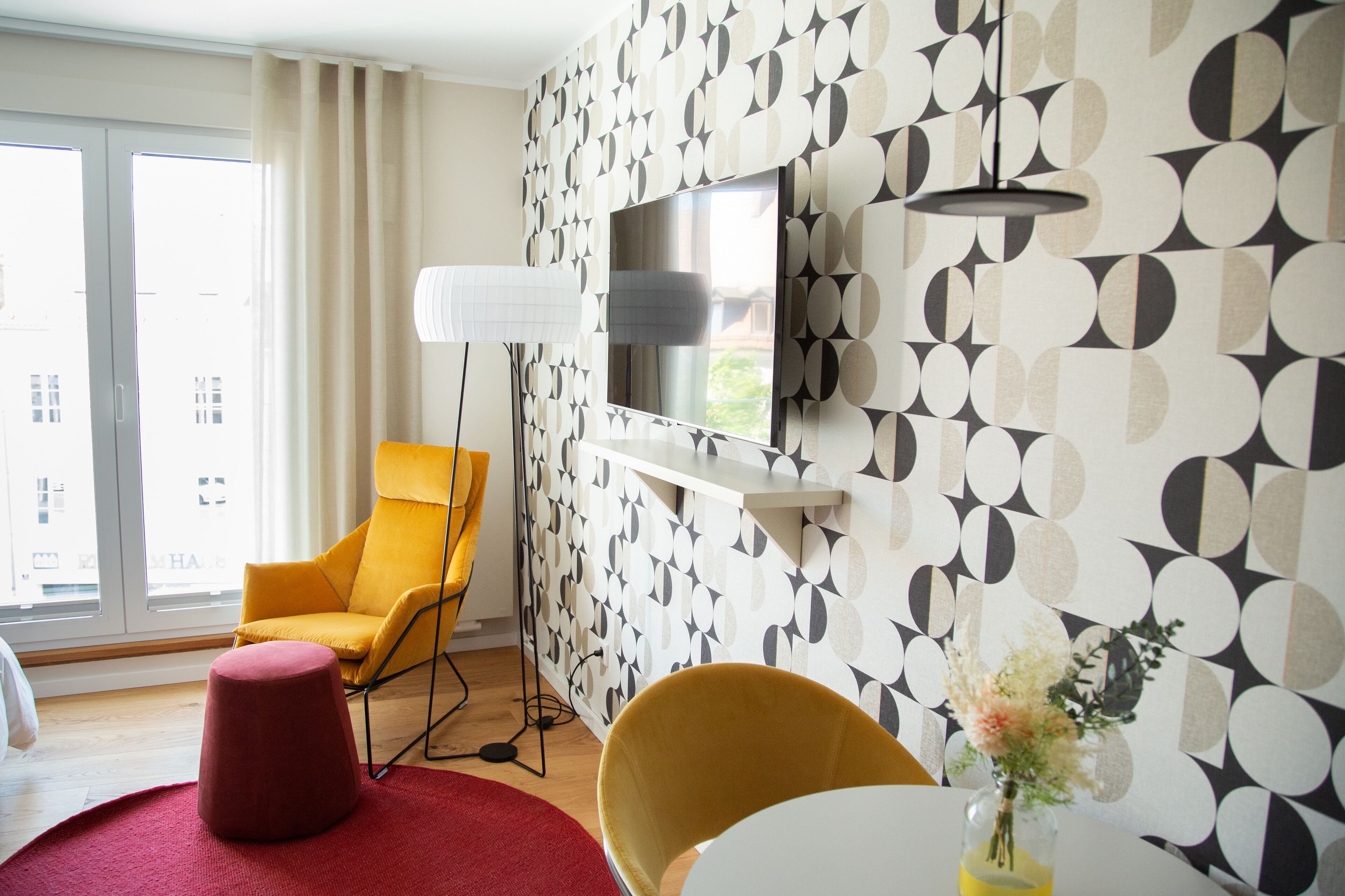 Ein modernes Zimmer mit an der Wand montiertem Fernseher, einem gelben Sessel, einem roten Hocker und einer Tischvase mit Blumen.