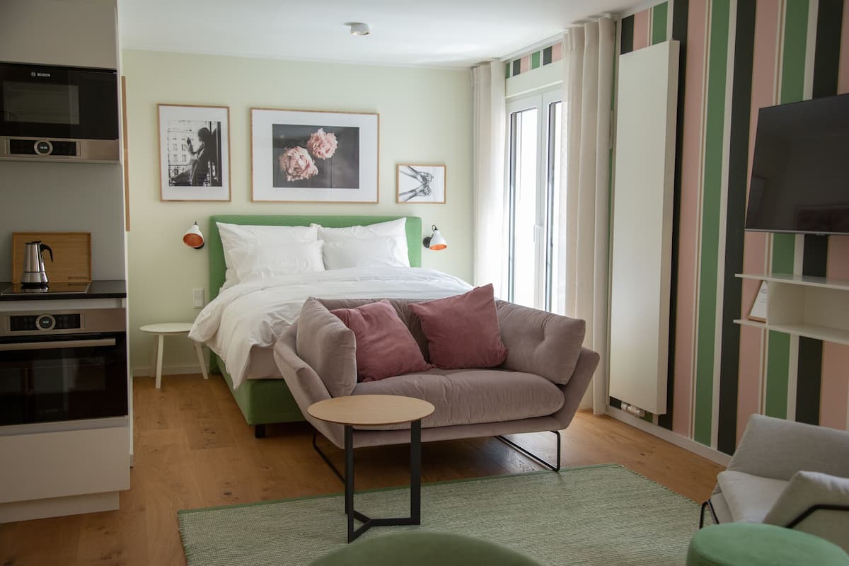 A modern bedroom with a bed, a green armchair, a small table, a TV, and a kitchenette.
