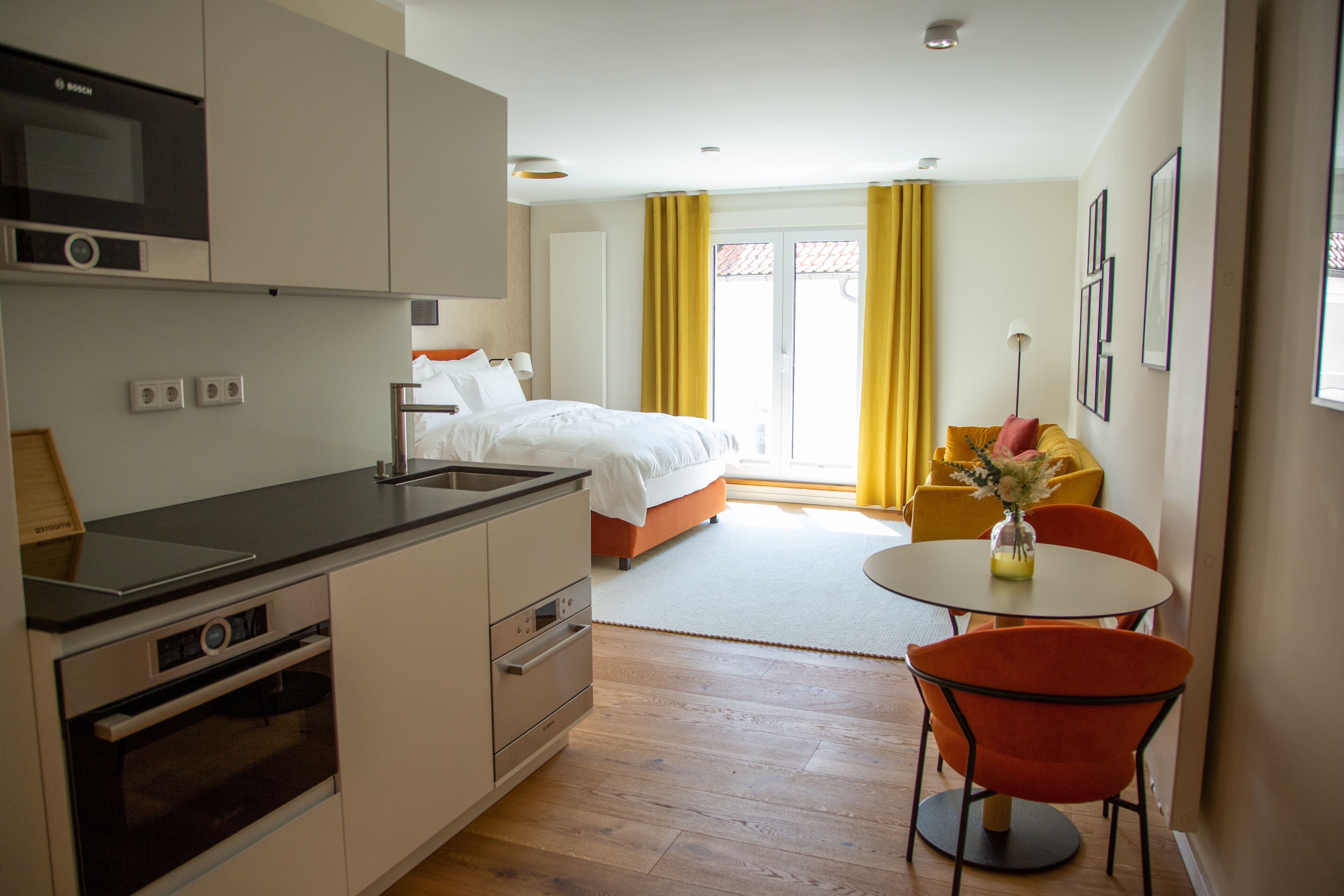 Una camera d'albergo moderna con angolo cottura, un letto, zona pranzo con tavolo e sedie, e un balcone con tende gialle.