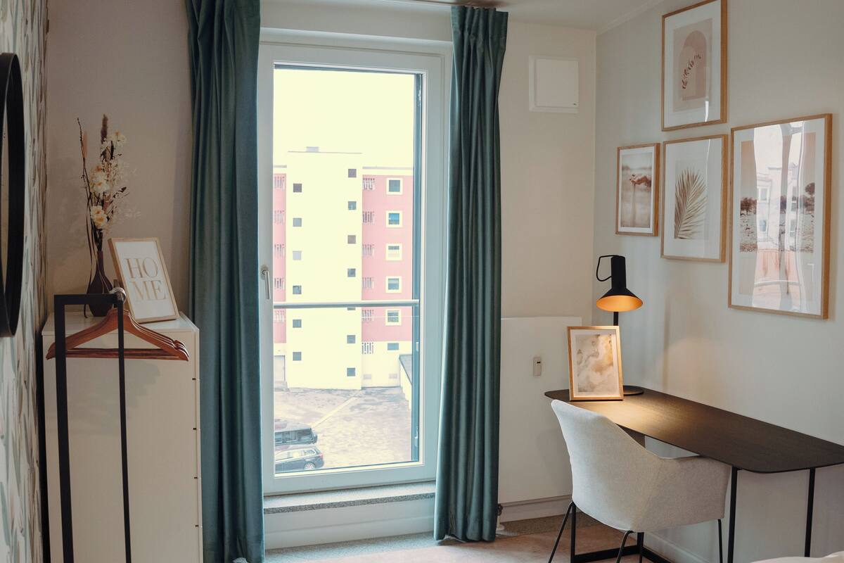 A room with a desk, chair, framed pictures, a window with curtains, and a view of buildings outside.