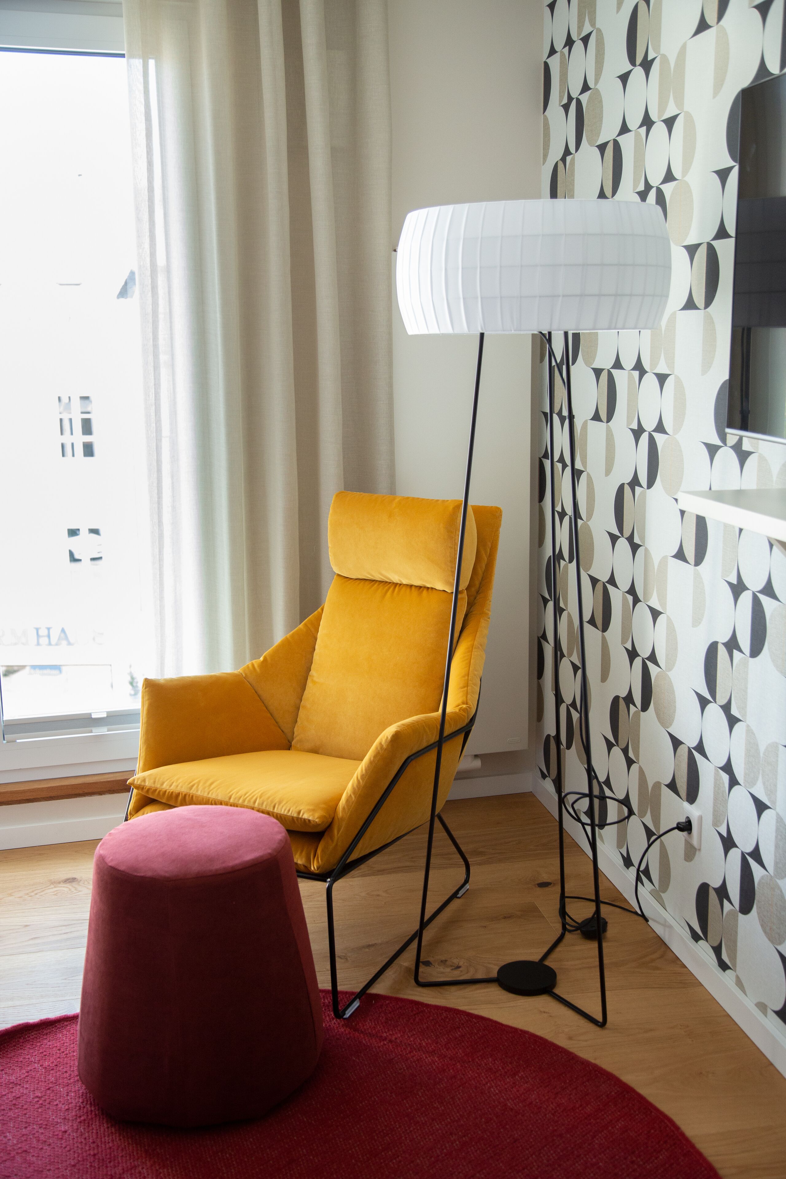 Una sala de estar moderna con un sillón amarillo, un otoman rojo, una lámpara de pie y una pared con estampado.