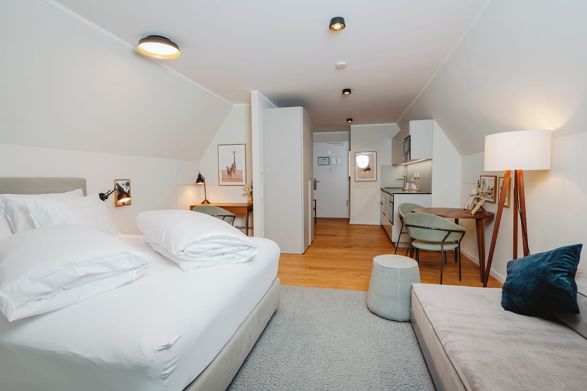 A modern hotel room with a large bed, a desk, a chair, and a small kitchenette.
