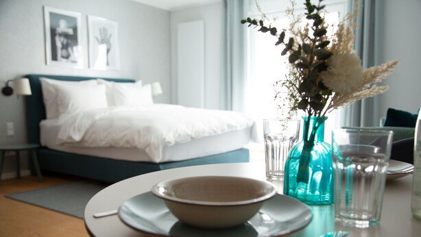 Uma área de refeições bem organizada, com uma mesa posta para dois, um vaso com flores e uma cama ao fundo.