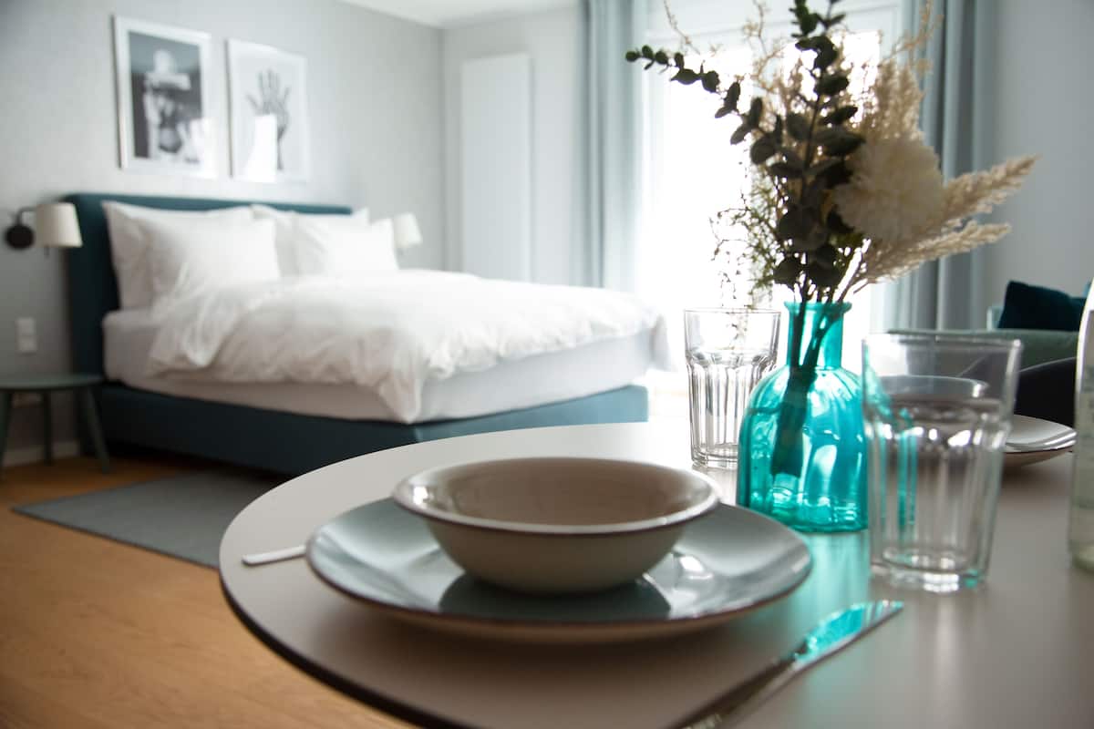 A neatly arranged dining area with a table set for two, a vase with flowers, and a bed in the background.