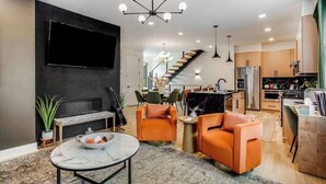 Ein modernes Wohnzimmer mit Flachbildfernseher, orangefarbenen Sesseln, einem Marmortisch und einer Treppe, die in einen Küchenbereich führt.