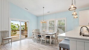 A dining area with a table and chairs, a kitchen island with a sink, and a view through large windows to the outside. - Tranquility Cove: New Build, Elevator, Pool Access (Nags Head)