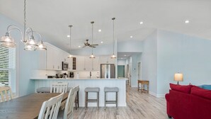 A modern kitchen with white cabinets, a central island with bar stools, and pendant lighting. - Tranquility Cove: New Build, Elevator, Pool Access (Nags Head)