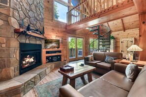 Interior acogedor de una cabaña con chimenea de piedra, muebles de madera, una escalera en espiral y grandes ventanales.