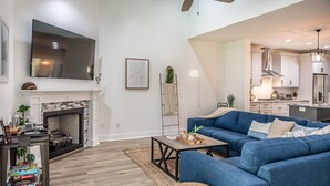Ein modernes Wohnzimmer mit Kamin, großem Flachbildfernseher, blauem Sofa und einer Küche im Hintergrund.