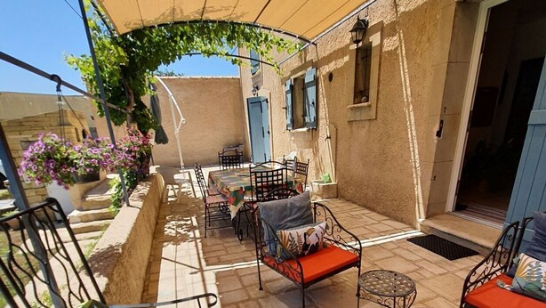 Een overdekt terras met tafels en stoelen, een pergola begroeid met klimplanten, en een stenen muur.