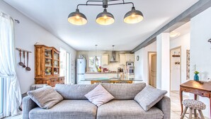A modern living room with a grey sofa, a glass coffee table, and a wooden dining table with a stool. There is a kitchen in the background with white cabinets and a hanging light fixture.