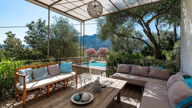 A covered outdoor seating area with a pool in the background, surrounded by trees and mountains.
