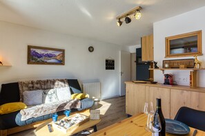 Una sala acogedora con sofá, mesa de centro y mesa de comedor con copas de vino y una botella. Se observa una cocina con armarios de madera y una ventana sobre la encimera.