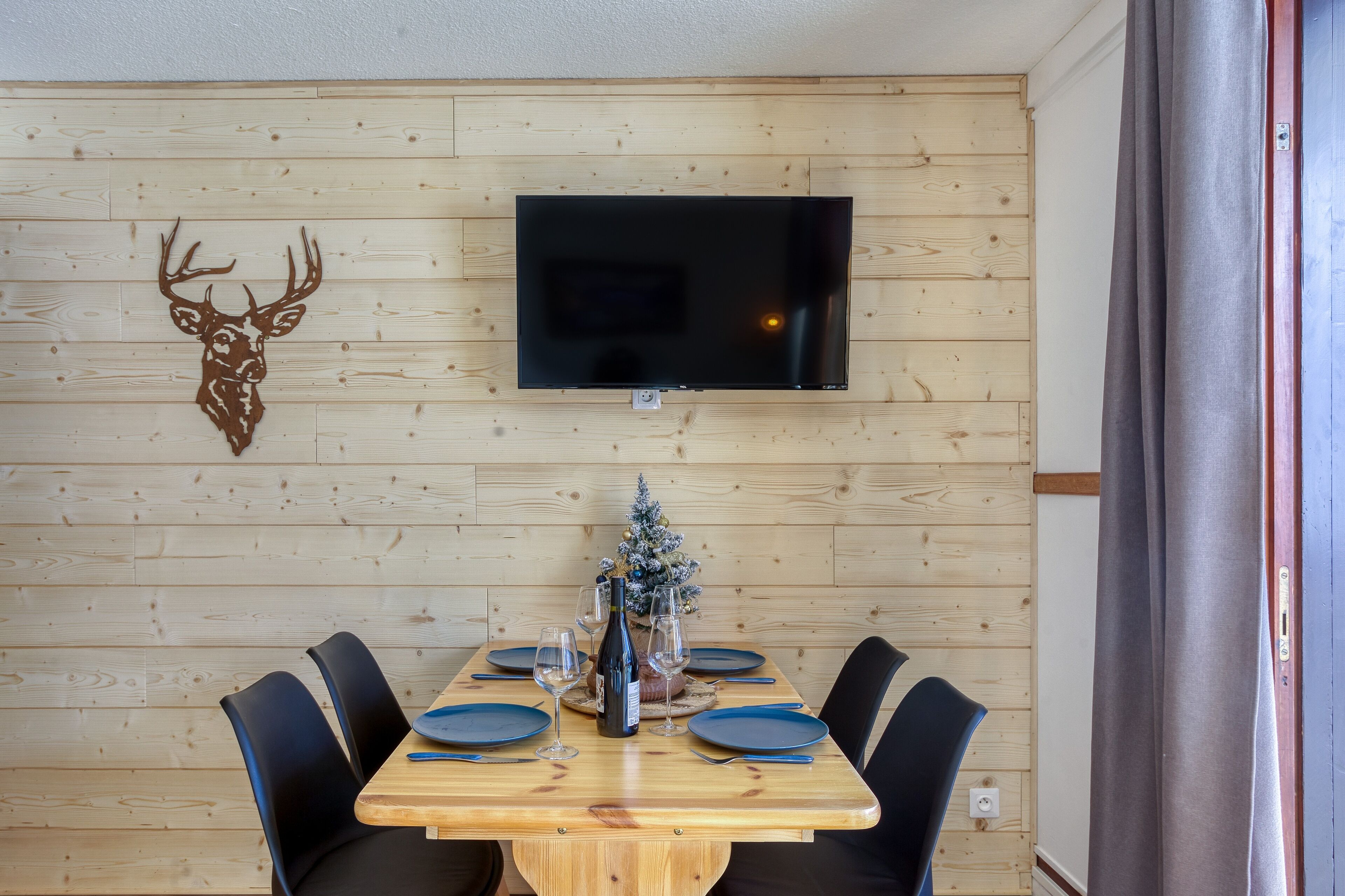 Un comedor con una mesa de madera, sillas, un televisor montado en la pared y un pequeño árbol de Navidad.