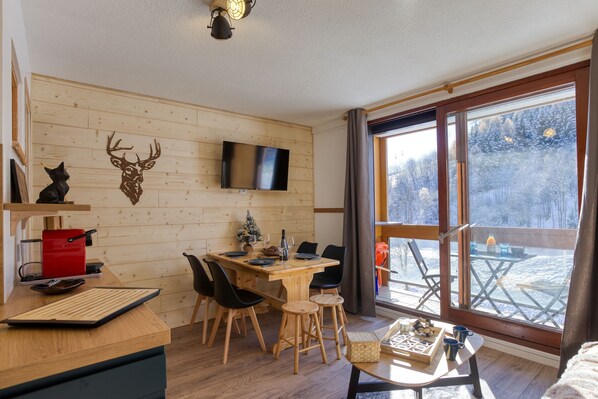 Un cuarto acogedor con pared de madera, un televisor, una mesa de comedor con sillas y un ventanal con vistas a un paisaje nevado.