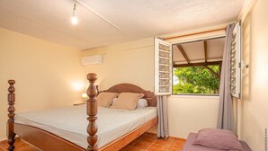 Een slaapkamer met een houten bed, een klein nachtkastje, een raam met gordijnen en uitzicht op groen.