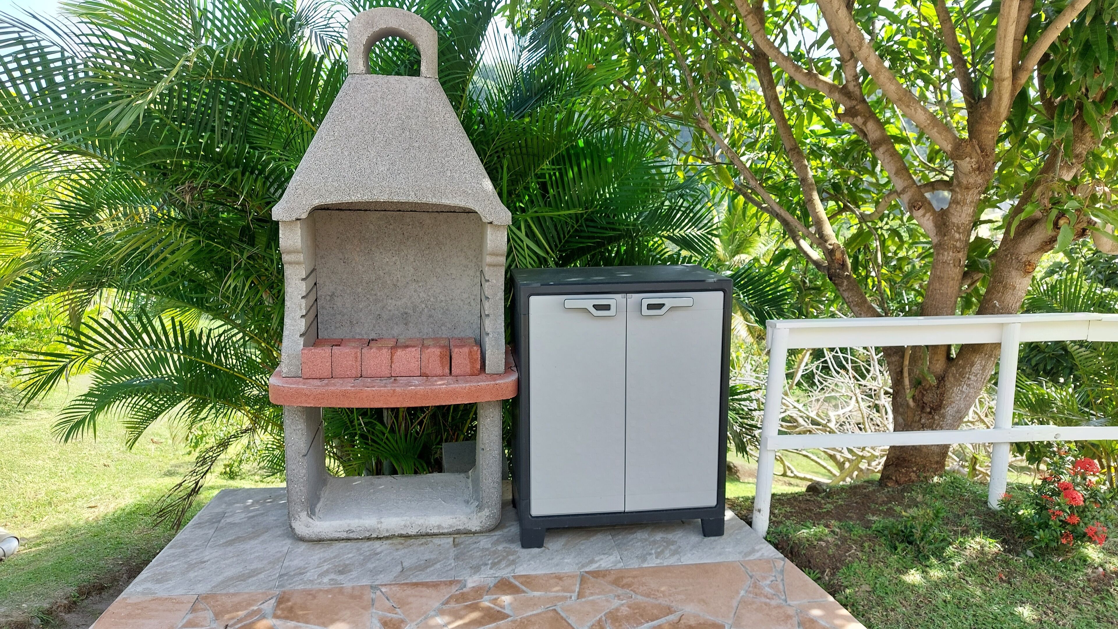 A stone-built outdoor grill with a small brick oven, a white metal cabinet, and a white fence in a lush garden.