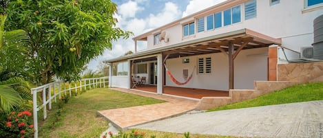 Une maison à deux étages dotée d’un patio couvert, d’une antenne parabolique et d’un jardin où poussent des fleurs rouges.