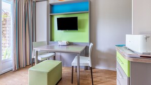 A modern room with a flat-screen TV mounted on a green wall, a wooden table with chairs, a green bench, and a kitchenette area with a microwave. - Danou - Appartement avec Terrasse (Courseulles-sur-Mer)