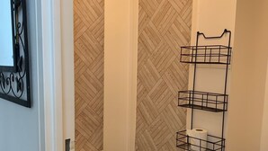 A bathroom with a white toilet, a shower with a herringbone-patterned wall, and a black metal shelf holding toilet paper.