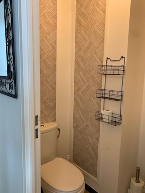 Une salle de bain avec des toilettes blanches, une douche dont le mur est décoré d’un motif en chevrons et une étagère en métal noir sur laquelle sont posés des rouleaux de papier toilette.