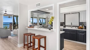 Una cocina moderna con armarios oscuros, una encimera blanca y vistas al océano. Hay dos taburetes de madera y un florero con flores sobre la encimera.