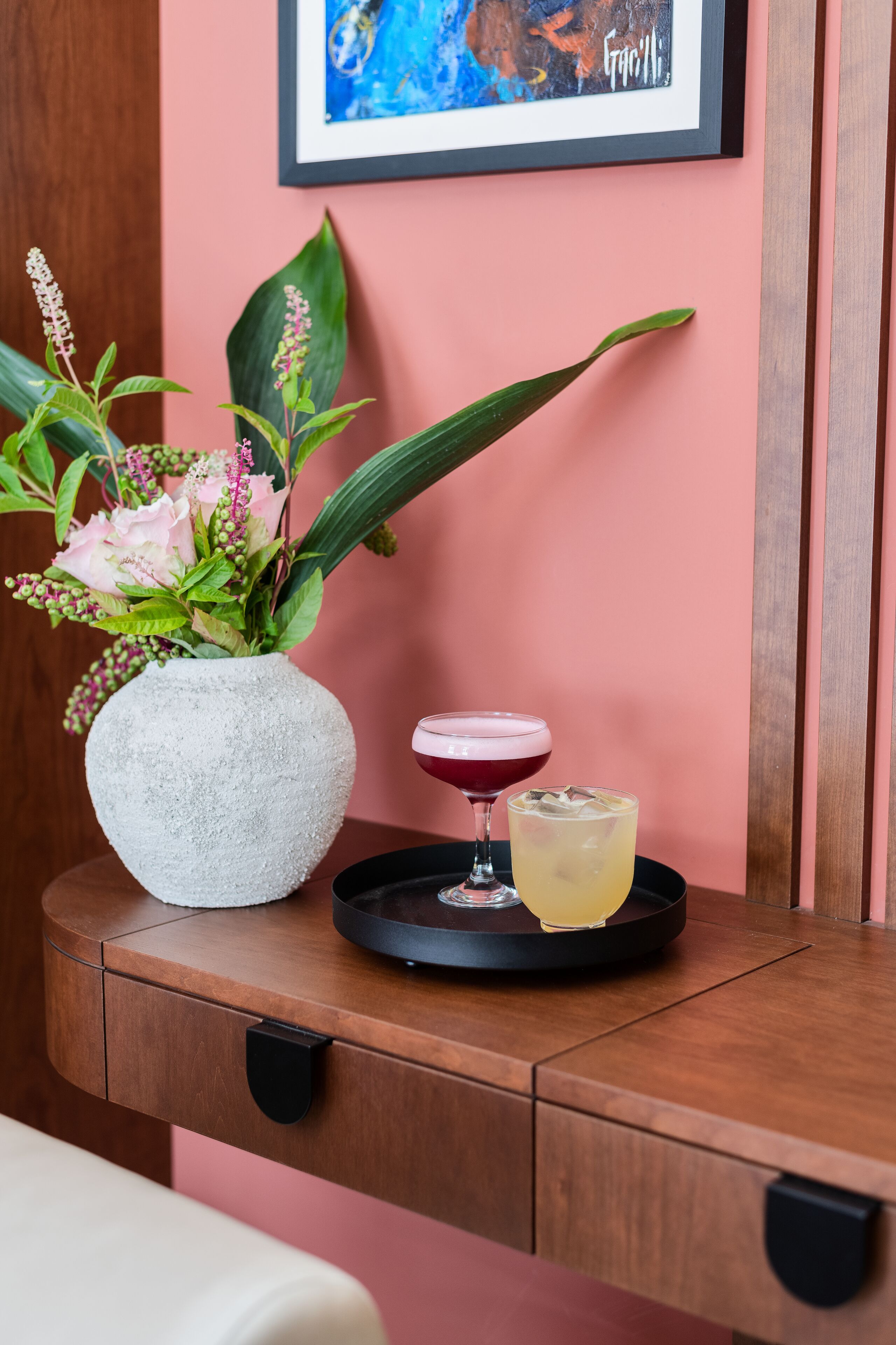 Una mesa de madera con un jarrón blanco lleno de flores y dos bebidas sobre una bandeja.