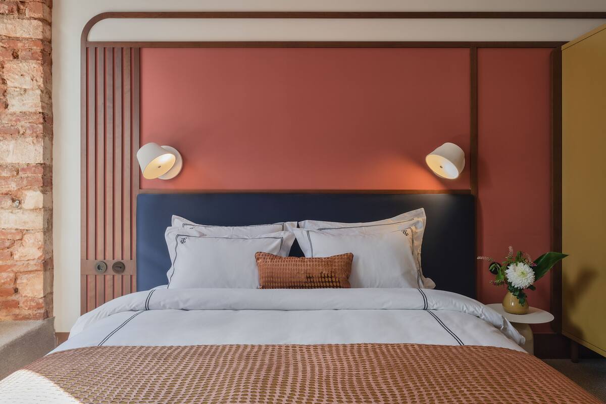 Un letto rifatto con lenzuola bianche e una coperta decorata, una testiera con linee verticali, due lampade a parete e un tavolino con un vaso di fiori.