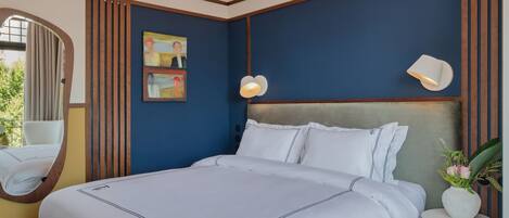 A neatly made bed with white linens and a wooden headboard, a round mirror, and a painting on the wall.