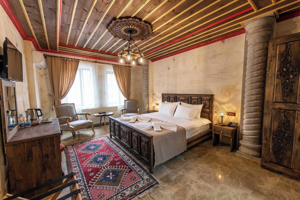 Quarto tradicional com teto de madeira, uma cama grande, uma escrivaninha, uma cadeira e um tapete estampado.