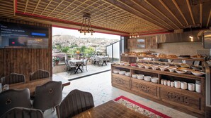 A restaurant with a wooden interior, a buffet counter with various dishes, and a view of a cityscape through large windows. - Volare Stone Hotel (Nevsehir)